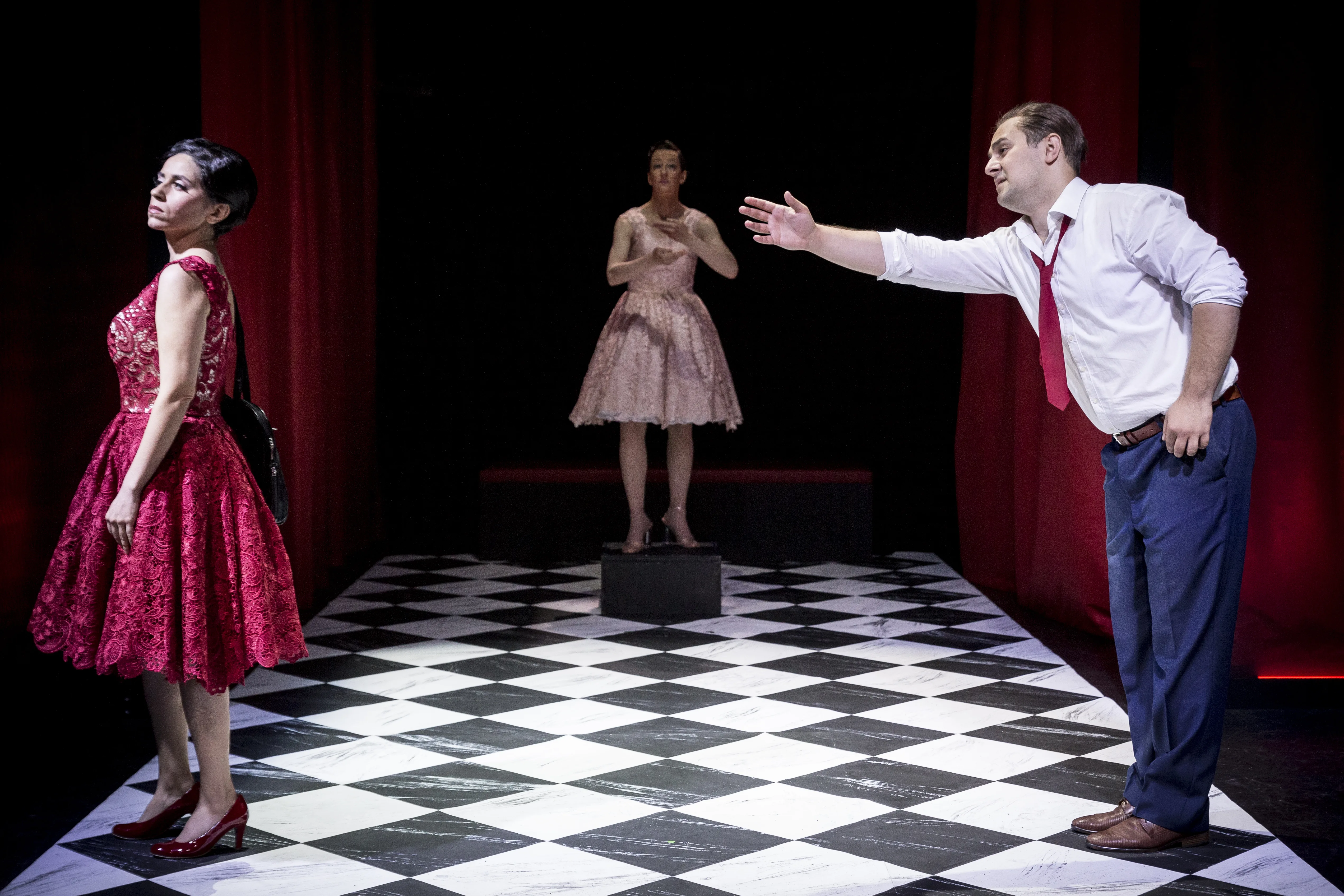 Das Bild zeigt eine theatrale Inszenierung mit drei Personen auf einem markanten schwarz-weiß gemusterten Karoboden, der an ein Schachbrett erinnert. Der Hintergrund ist dunkel gehalten und wird von schweren, roten Vorhängen an den Seiten eingerahmt.