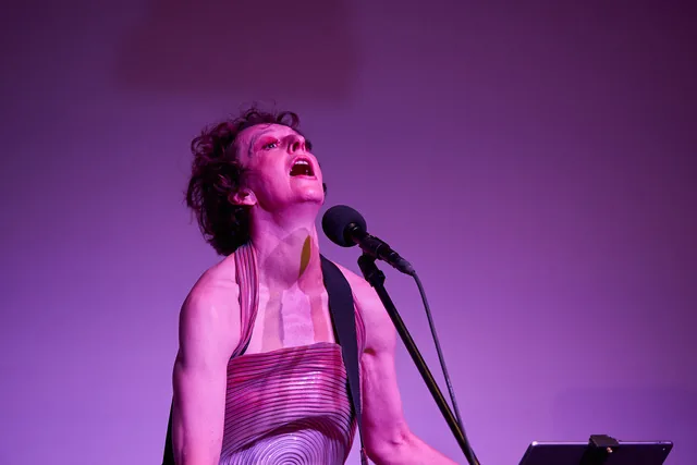 Gina Mattiello tritt auf einer Bühne vor einem violetten Hintergrund auf. Sie trägt ein markantes, strukturiertes Kleid mit konzentrischen Kreisen und singt in ein Mikrofon, während sie ein Instrument spielt. Auf einem Ständer vor ihr befindet sich ein Tablet.