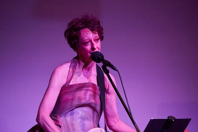 Gina Mattiello tritt auf einer Bühne vor einem violetten Hintergrund auf. Sie trägt ein markantes, strukturiertes Kleid mit konzentrischen Kreisen und singt in ein Mikrofon, während sie ein Instrument spielt. Auf einem Ständer vor ihr befindet sich ein Tablet. Zweites Bild