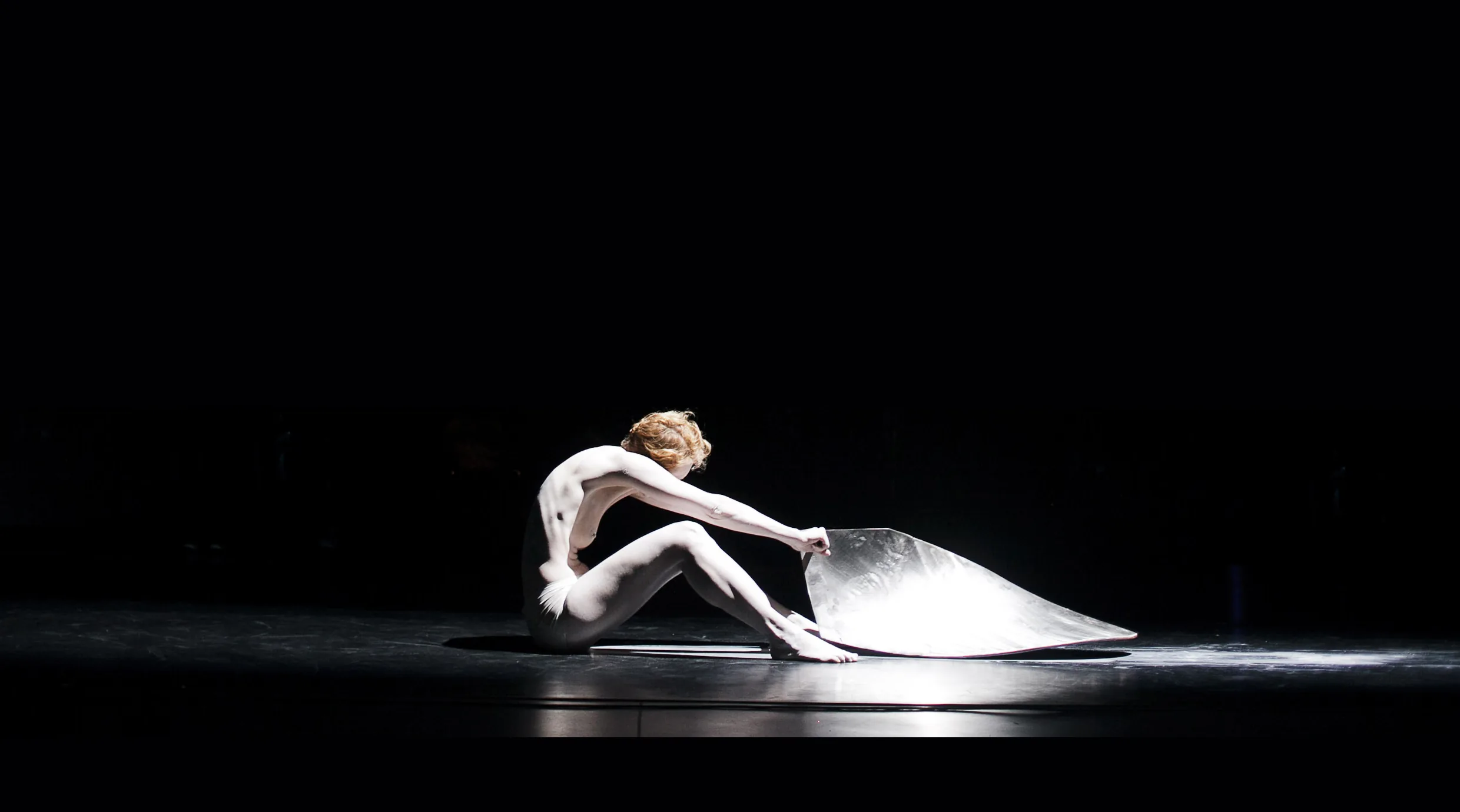 Eine minimalistische Theater-Performance auf dunkler Bühne. Gina Mattiello sitzt im Profil im Scheinwerferlicht auf dem Boden und interagiert mit einem großen, reflektierenden Metallelement, das sich vor ihr wölbt.