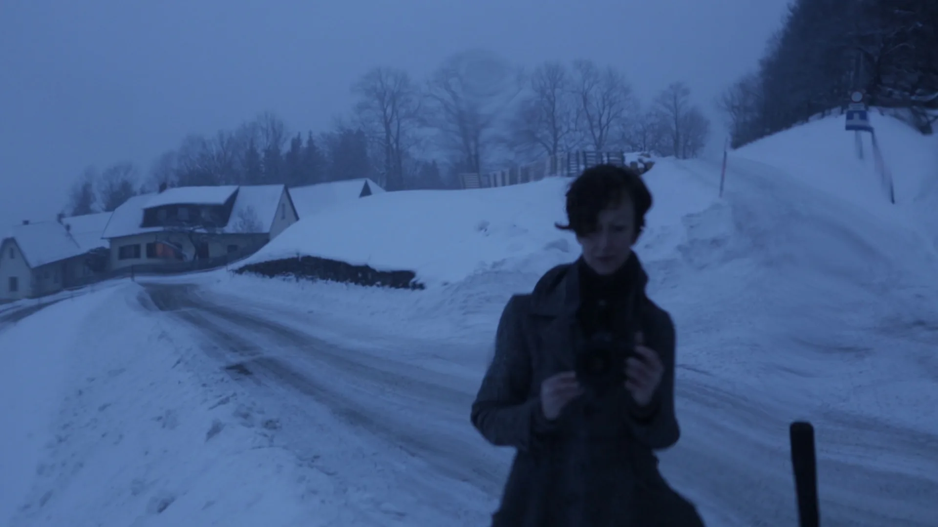 Eine dämmerige, blau getönte Winterlandschaft mit tief verschneiten Häusern und einer Straße. Im Vordergrund steht die Silhouette von Gina- sie hält eine Kamera, während die Umgebung in eine kühle, neblige Atmosphäre getaucht ist.