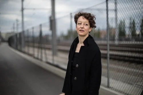 Porträtaufnahme von Gina mit kurzen, lockigen braunen Haaren und einem schwarzen Mantel. Sie blickt nachdenklich zur Seite; im Hintergrund erstreckt sich unscharf ein langer Maschendrahtzaun entlang von Bahngleisen unter bewölktem Himmel.