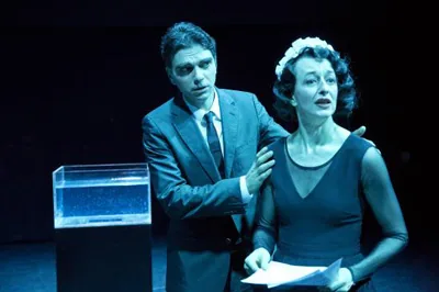Szenenfoto einer Theateraufführung in kühlem Blaulicht. Ein Mann im Anzug legt einer Frau im dunklen Kleid beruhigend die Hand auf die Schulter. Die Frau hält ein Skript in der Hand; links im Bild steht ein gläsernes, mit Wasser gefülltes Gefäß auf einer Säule.