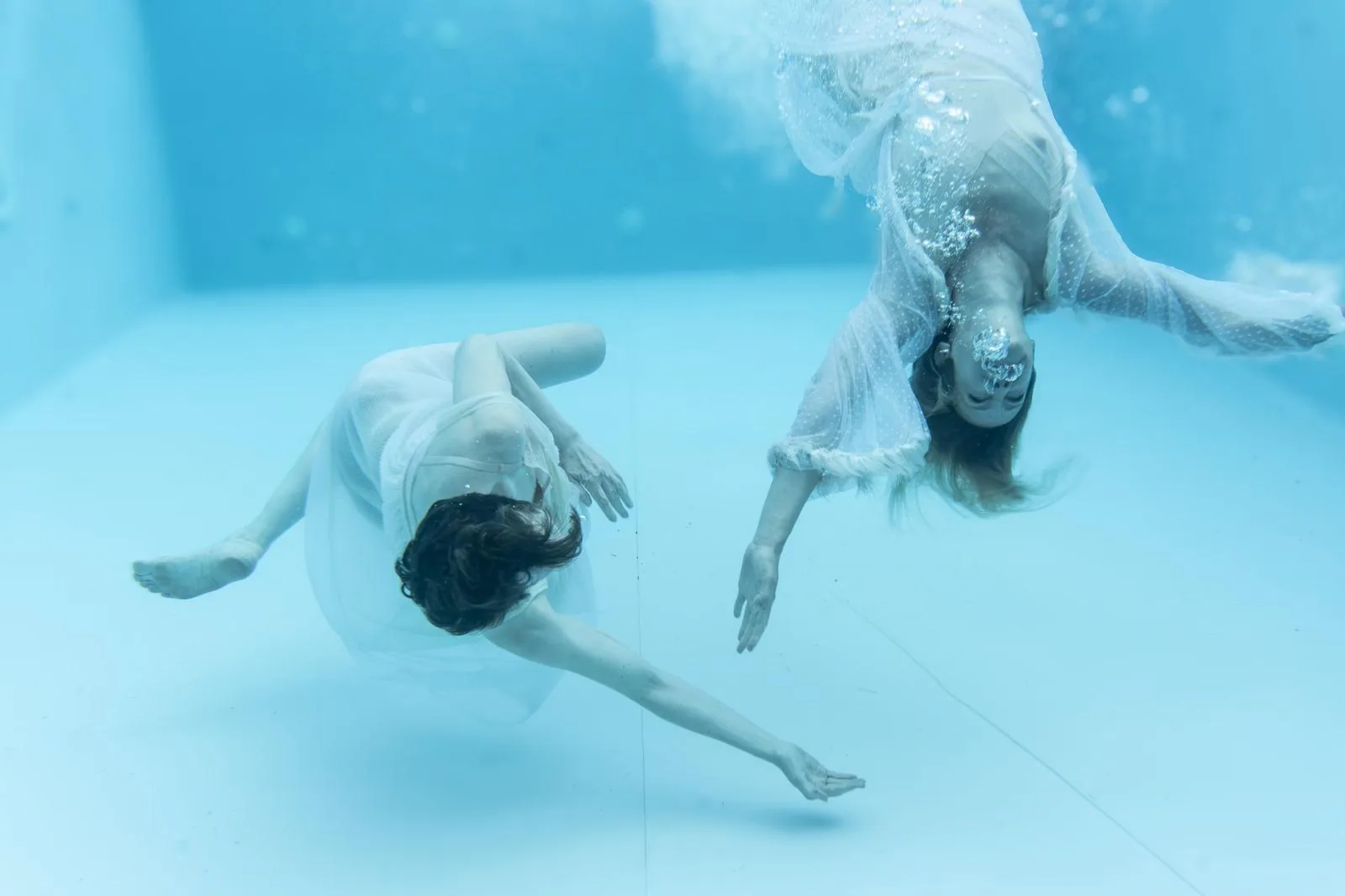 Zwei Frauen in fließenden, weißen Kleidern schweben in einer surrealen Unterwasserszene in hellem, blauem Wasser. Ihre Bewegungen wirken schwerelos und tänzerisch, während kleine Luftblasen zur Oberfläche steigen.